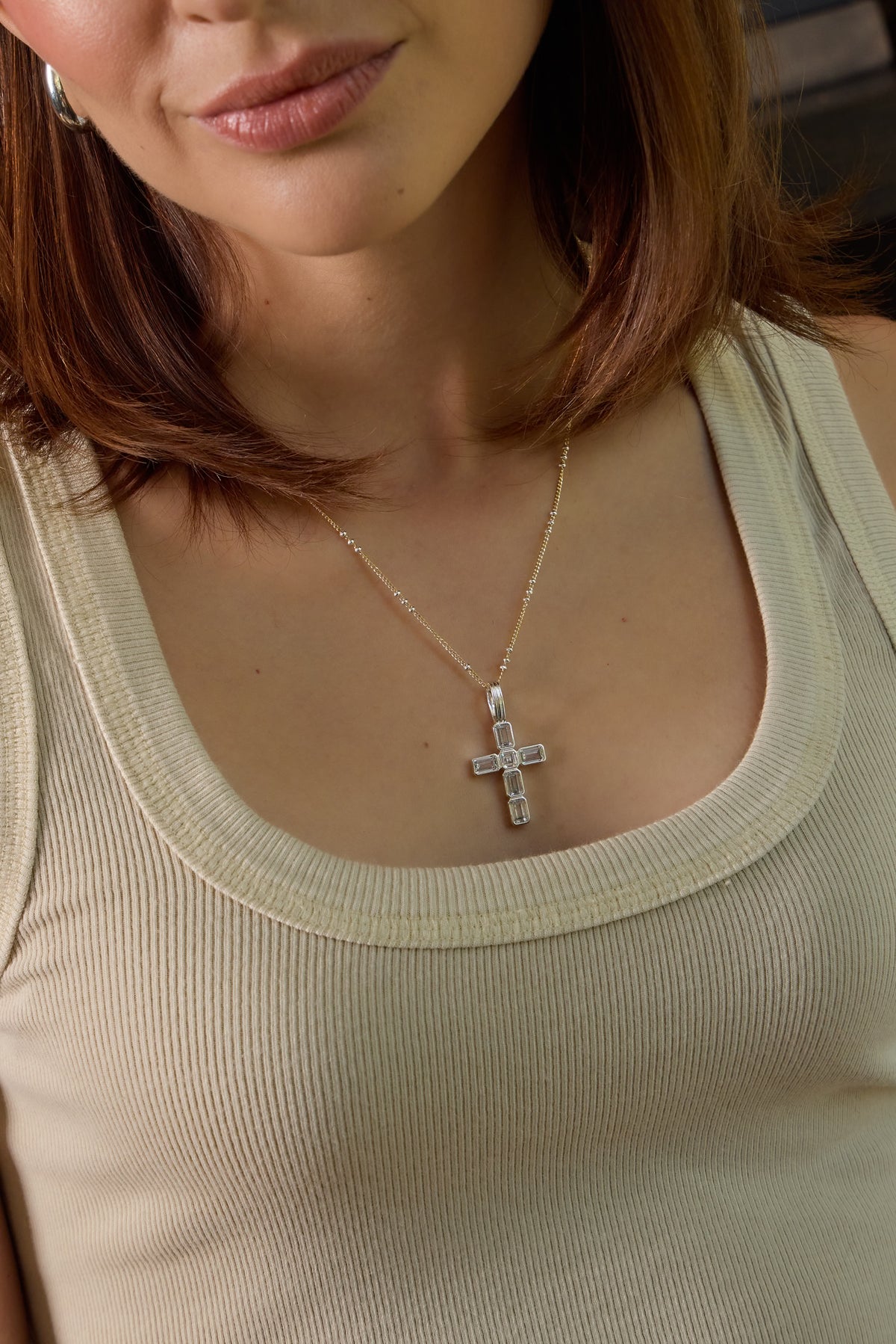 A close-up of a woman wearing the Grace silver cross charm with clear emerald CZ's styled with a beige tank top.