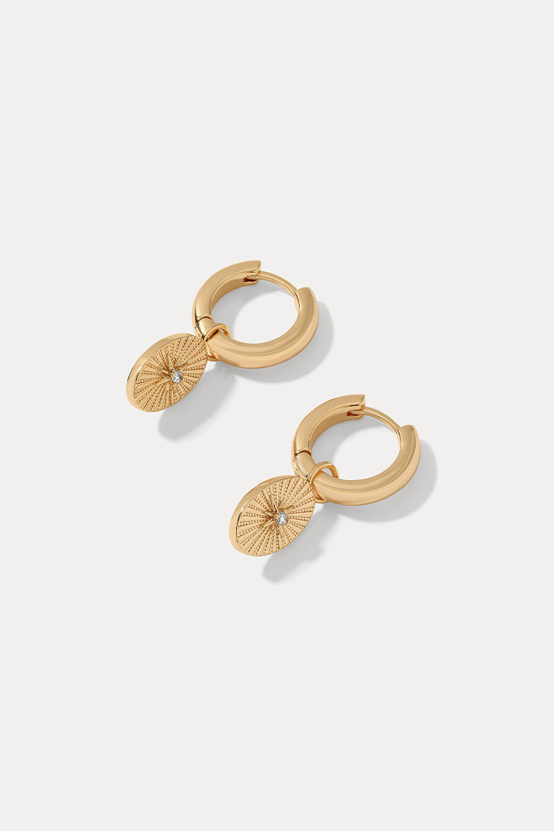 A pair of gold huggie hoop earrings with textured sunburst oval drops, each featuring a small clear crystal in the center.