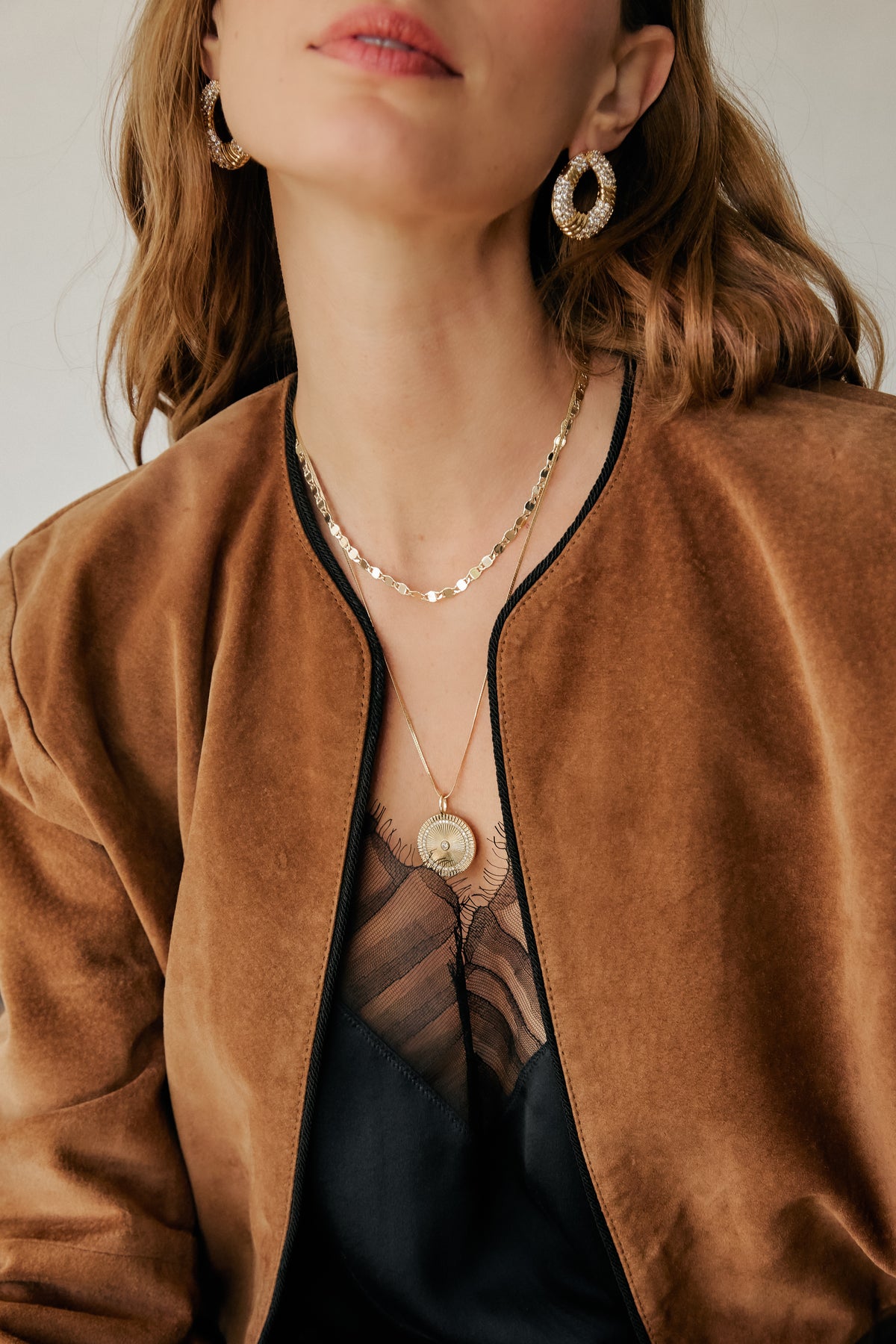 Model wearing layered gold Ava necklace, Solara charm pendant, Halle earrings, and brown jacket.