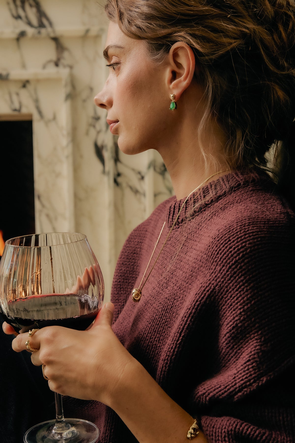 Model in burgundy sweater holding wine glass, wearing Panther Emerald drop earring, Celeste Slider, and Panther ring.